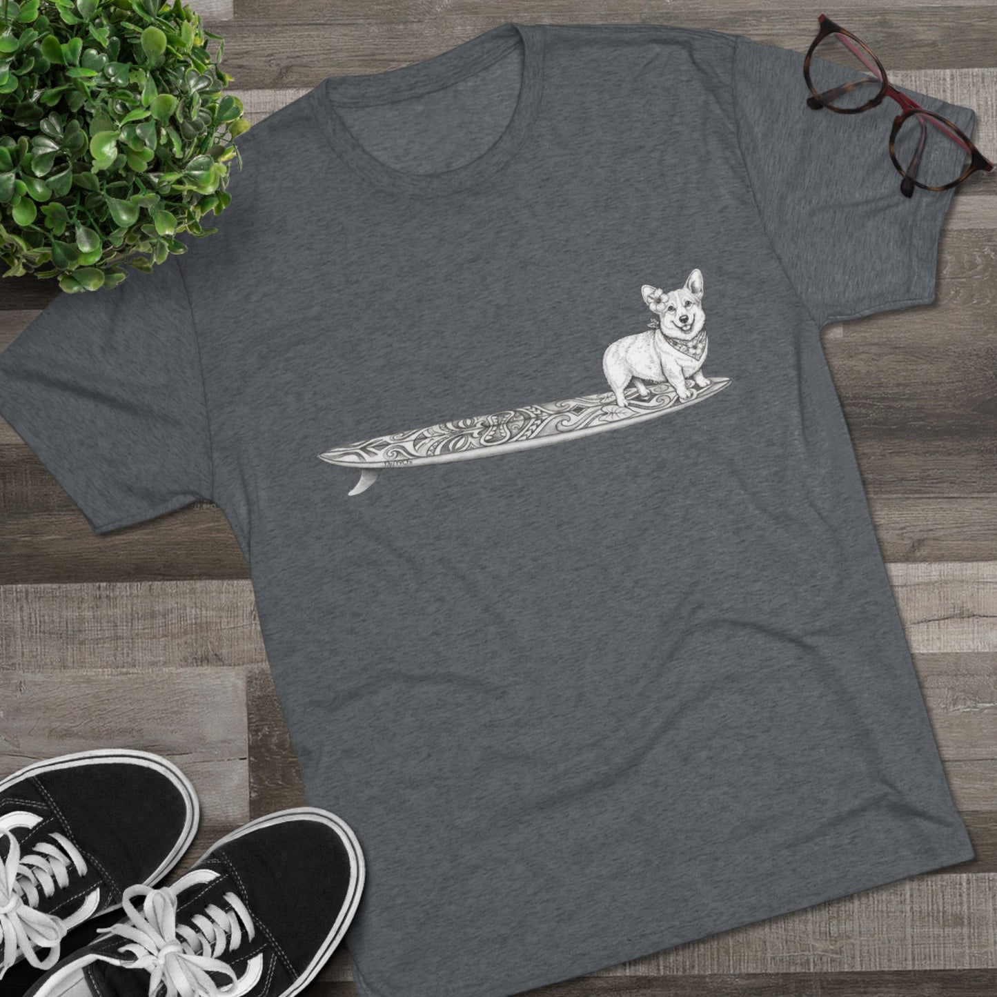 Graytshirt with a Corgi on a tiki art longboard with a hibiscus in it's ear and a hibiscus bandana around it's neck on a gray background.