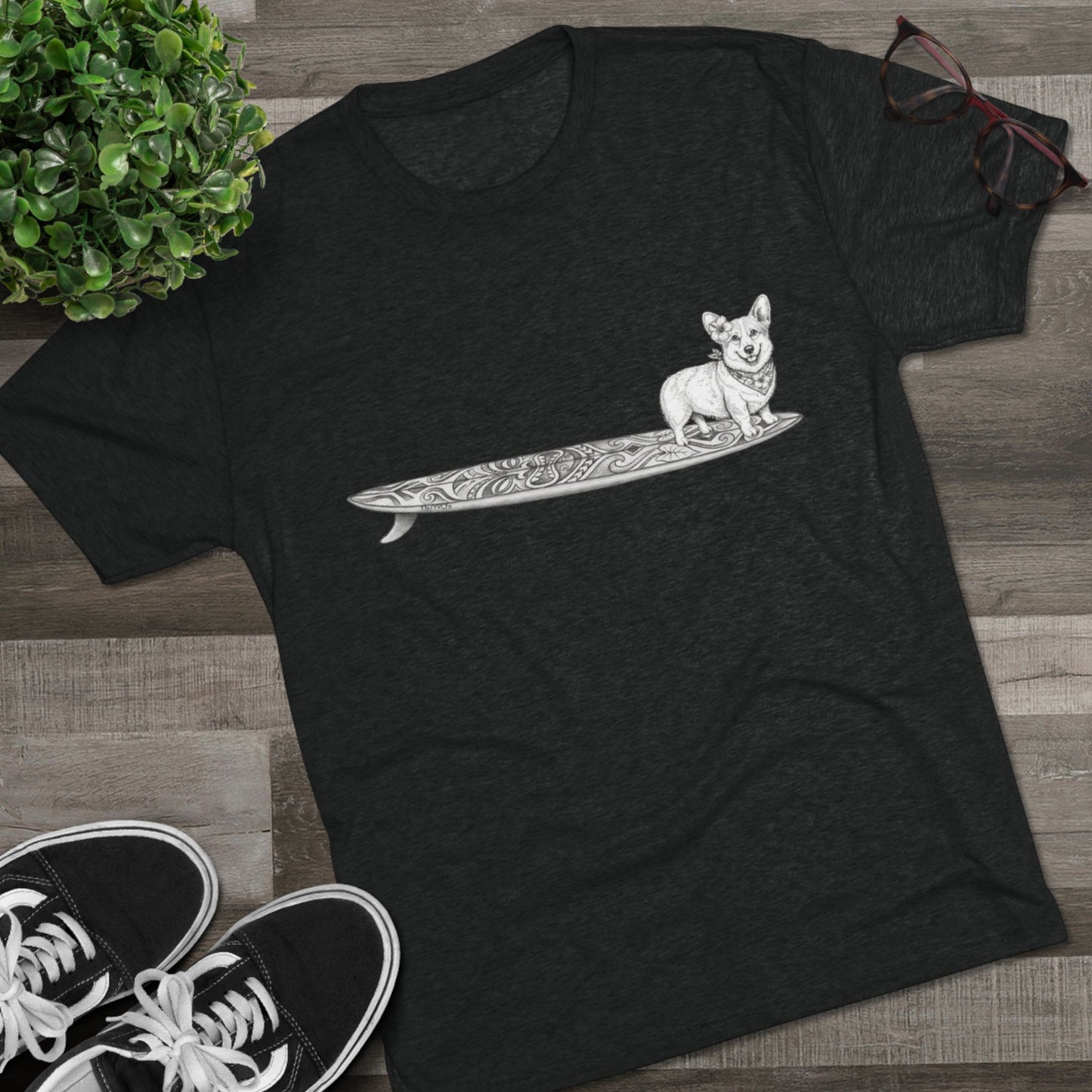 Black tshirt with a Corgi on a tiki art longboard with a hibiscus in it's ear and a hibiscus bandana around it's neck on a black background.