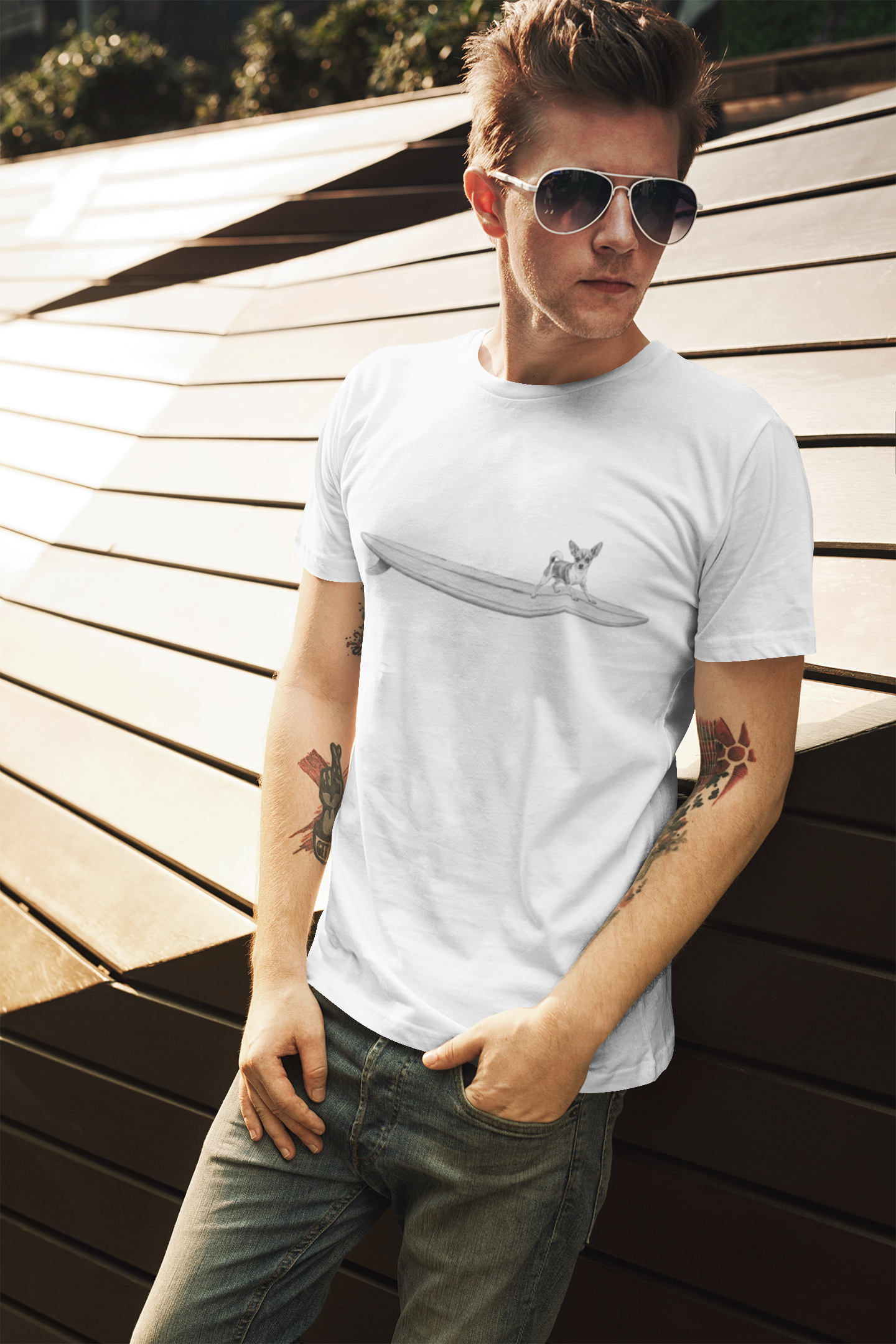Man wearing a white t-shirt with a graphic of a Chihuahua on a longboard surfboard on a white background wearing sunglasses, and tattoos standing against a wooden background.