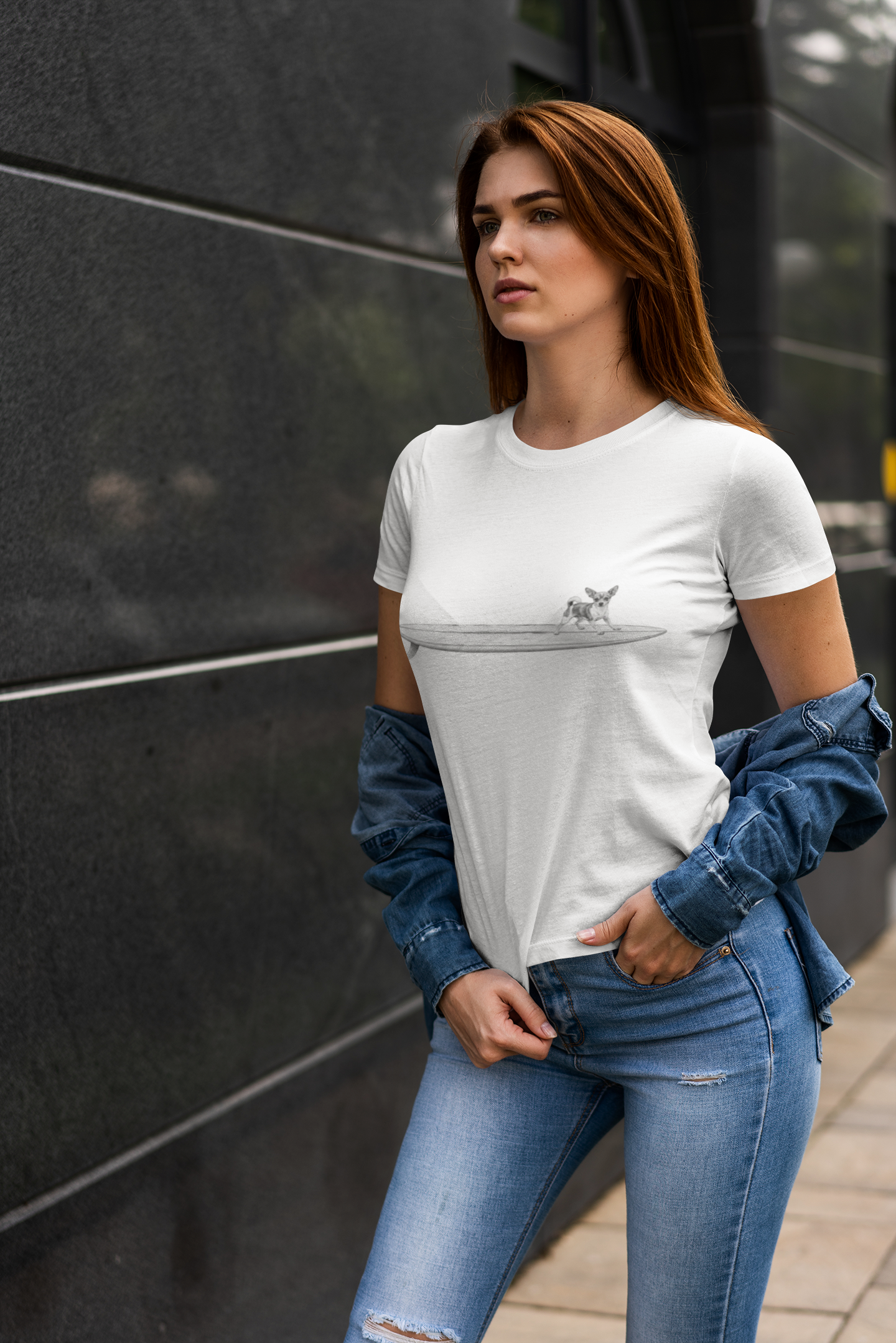 Woman wearing a white t-shirt with a graphic of a Chihuahua on a longboard surfboard on a white background in blue jeans against a dark wall.