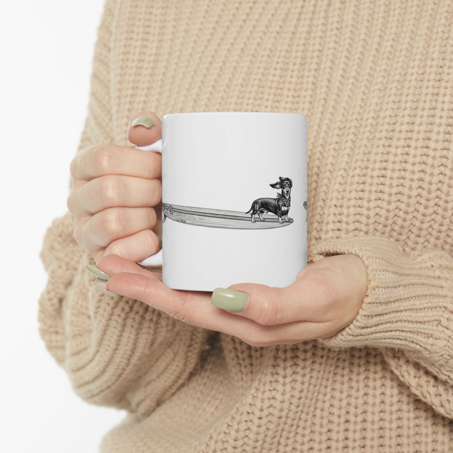 Person holding a White mug with a black and white illustration of a white mug with a dachshund with a checkered scarf hanging twenty on a surfboard graphic with checkered racing flags on a white background mug with a dachshund illustration. person is wearing a beige sweater.